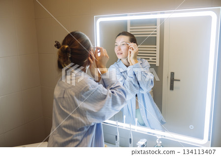 Woman applying eye cream at LED mirror - evening skincare ritual 134113407