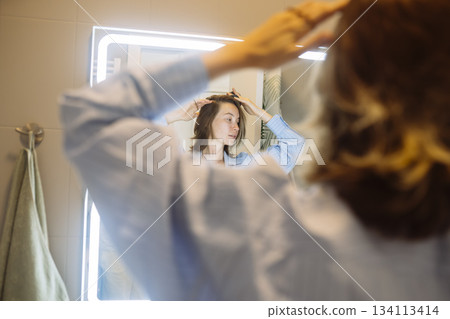 woman checking hair in illuminated mirror, morning routine 134113414