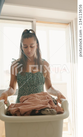 A woman is skillfully carrying a laundry basket filled with clothes in a bright interior space A woman is skillfully carrying a laundry basket filled with clothes in a bright interior space 134113656