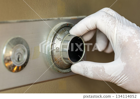 Gloved hand adjusting the dial on an old combination safe lock with visible wear on metal and beige safe body 134114052