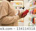 African American woman compares two bottles of haircare products in a store, making a conscious choice while shopping for shampoo or cosmetic treatment. 134114318