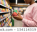 African American woman choosing canned dog food at a pet store, reading the label. Customer compares wet pet food in the aisle with various canned goods on the shelves Concept of responsible pet owner 134114319