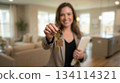 Close-up of keys with blurred smiling woman real estate agent offering new home to client, holding documents, standing in modern bright apartment interior. 134114321