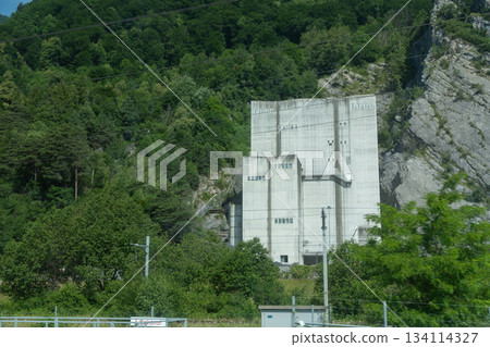 A concrete industrial structure set against a green mountain slope. Dense trees, rocky terrain, and the solid geometric building make the scene suitable for themes of industry, infrastructure, and 134114327