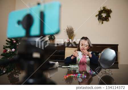 Online creator Caucasian child girl talks about her favorite beauty products. Livestream filmed using smart mobile phone on tripod at home. 134114372