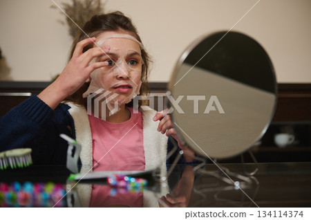 Elementary age Caucasian girl applying moisturizing face mask, siting at a table and admiring her appearance in a cosmetic mirror. The concept of home spa and skin care Elementary age Caucasian girl applying moisturizing face mask, siting at a table and admiring her appearance in a cosmetic mirror. The concept of home spa and skin care 134114374