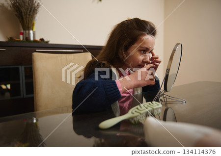 Beautiful child girl applying moisturizing face mask, siting at a table and admiring her appearance in cosmetic mirror 134114376