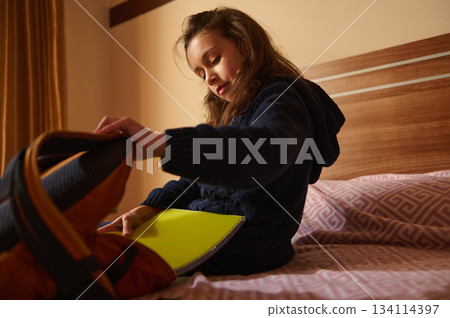 Cute elementary age school child girl arranging her backpack, putting copybooks inside it while sitting on her bed at home, preparing for going to school Cute elementary age school child girl arranging her backpack, putting copybooks inside it while sitting on her bed at home, preparing for going to school 134114397