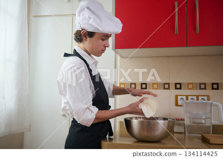 Teenage Boy Preparing Pizza at Home Kitchen in Black Apron and White Chef Hat Teenage Boy Preparing Pizza at Home Kitchen in Black Apron and White Chef Hat 134114425