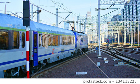 Warsaw, Poland. 28 November 2025. A passenger train on the tracks near the station. Tracks, electrical wires and metal masts against the backdrop of a city skyscrapers on a winter day.  134115223