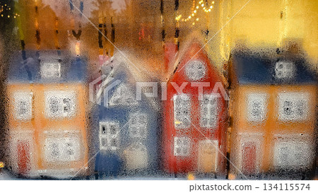 Blurred view of colorful miniature houses through a window covered in condensation, with warm holiday lights Blurred view of colorful miniature houses through a window covered in condensation, with warm holiday lights 134115574
