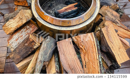 Close up of a warm metallic fire pit surrounded by chopped firewood and wet wooden deck boards 134115599