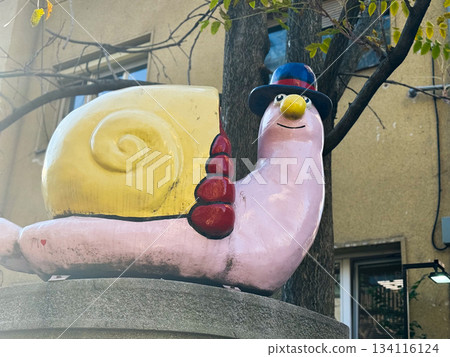 Whimsical snail sculpture with hat beside urban building and tree. Playful outdoor art, colorful street decoration, nostalgic charm and cheerful city atmosphere. Whimsical snail sculpture with hat beside urban building and tree. Playful outdoor art, colorful street decoration, nostalgic charm and cheerful city atmosphere. 134116124