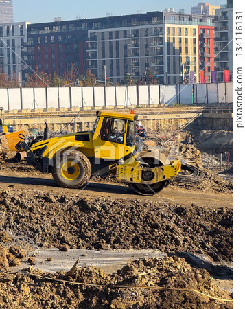 Yellow soil roller at construction site with high-rise buildings in background. Urban development, roadwork machinery, infrastructure improvement and foundation compaction process. 134116131