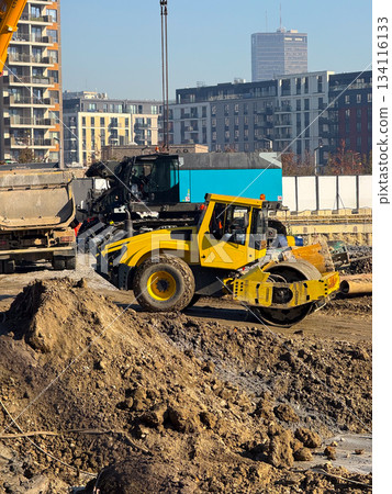 Yellow soil roller at construction site with high-rise buildings in background. Urban development, roadwork machinery, infrastructure improvement and foundation compaction process. 134116133