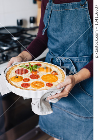 Female hands holding cheese and tomato quiche in the ceramic baking dish. 134116637