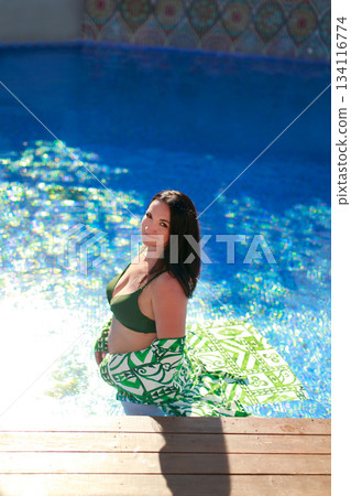 Pregnant woman relaxing in swimming pool wearing green bikini and stylish beach dress Pregnant woman relaxing in swimming pool wearing green bikini and stylish beach dress 134116774