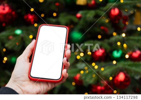 Woman hand with mobile phone and empty white blank screen against Christmas tree. Screen display for design. Xmas Holiday Mock up. Christmas advertising, smartphone app template Woman hand with mobile phone and empty white blank screen against Christmas tree. Screen display for design. Xmas Holiday Mock up. Christmas advertising, smartphone app template 134117298