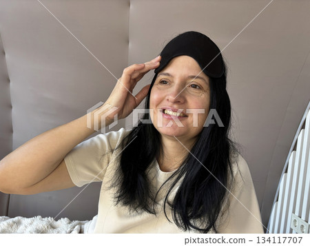 Adult brunette woman sitting in bed with a sleep mask, smiling and looking relaxed after a good night sleep, cozy morning routine, healthy sleep, wellness and self care lifestyle 134117707
