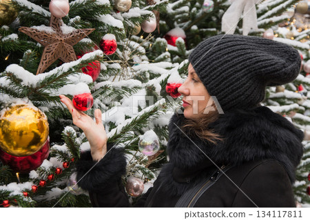 Woman standing near decorated Christmas fir tree with lights and ornaments at an outdoor festive market. 134117811
