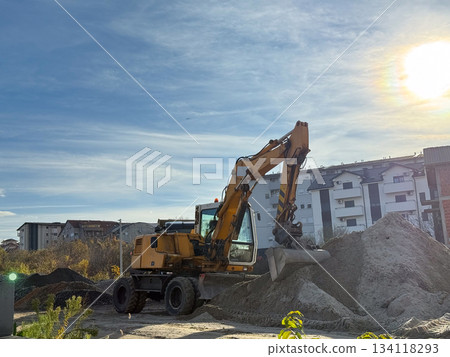 Yellow excavator digging sand on construction site under bright sun. Industry, machinery, earthmoving, development and urban building progress. Yellow excavator digging sand on construction site under bright sun. Industry, machinery, earthmoving, development and urban building progress. 134118293