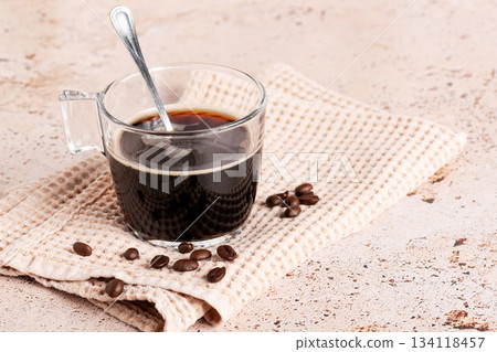 Hot black coffee in a clear glass cup with coffee beans on textured cloth Hot black coffee in a clear glass cup with coffee beans on textured cloth 134118457
