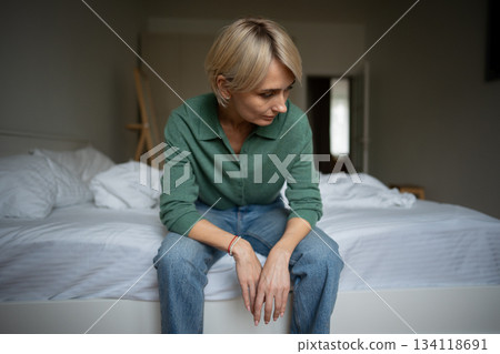 Pondering middle aged woman sits on bed in bedroom with uninterested expression, apathy and fatigue. 134118691
