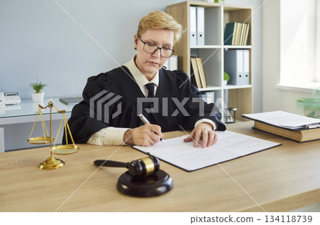 Female judge signing legal documents at courtroom desk with gavel and scales Female judge signing legal documents at courtroom desk with gavel and scales 134118739