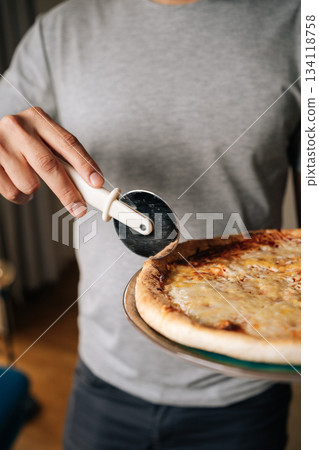 Vertical cropped shot of unrecognizable man wearing gray t-shirt holding round pizza on plate, cutting pizza using cutter wheel tool, ready to serve delicious slices for sharing at home, closeup. Vertical cropped shot of unrecognizable man wearing gray t-shirt holding round pizza on plate, cutting pizza using cutter wheel tool, ready to serve delicious slices for sharing at home, closeup. 134118758