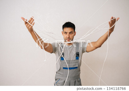 Puzzled electrician male wearing grey uniform holding bunch of tangled white electrical wires, looking confused and frustrated by complexity of wiring. Concept of wiring maintenance and service. Puzzled electrician male wearing grey uniform holding bunch of tangled white electrical wires, looking confused and frustrated by complexity of wiring. Concept of wiring maintenance and service. 134118766