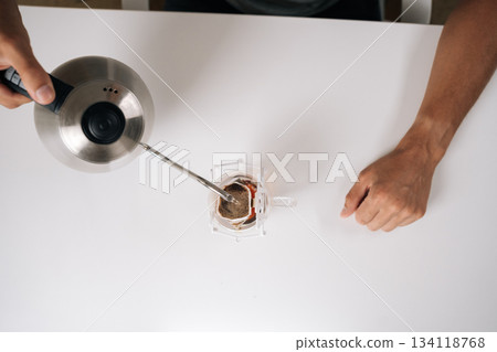 Top view of coffee lover brewing drip coffee with kettle, carefully pouring hot water over paper filter resting on transparent glass cup, all set on clean white table in cozy kitchen. Top view of coffee lover brewing drip coffee with kettle, carefully pouring hot water over paper filter resting on transparent glass cup, all set on clean white table in cozy kitchen. 134118768