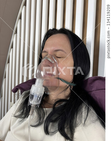 Adult woman using a home nebulizer inhaler while sitting in a cozy bedroom, respiratory treatment at home for breathing relief, healthcare and medical therapy concept, realistic lifestyle scene 134119209