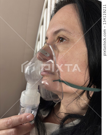 Adult woman using a home nebulizer inhaler while sitting in a cozy bedroom, respiratory treatment at home for breathing relief, healthcare and medical therapy concept, realistic lifestyle scene Adult woman using a home nebulizer inhaler while sitting in a cozy bedroom, respiratory treatment at home for breathing relief, healthcare and medical therapy concept, realistic lifestyle scene 134119211