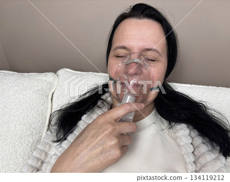 Adult woman using a home nebulizer inhaler while sitting in a cozy bedroom, respiratory treatment at home for breathing relief, healthcare and medical therapy concept, realistic lifestyle scene Adult woman using a home nebulizer inhaler while sitting in a cozy bedroom, respiratory treatment at home for breathing relief, healthcare and medical therapy concept, realistic lifestyle scene 134119212