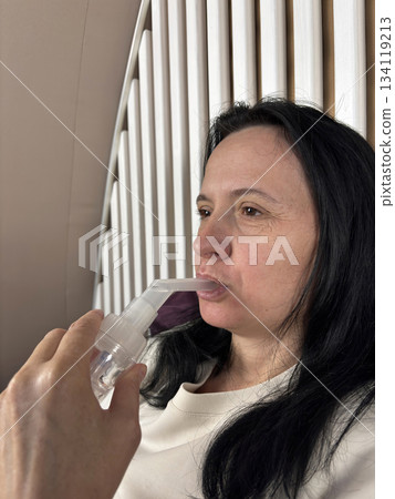 Adult woman using a home nebulizer inhaler while sitting in a cozy bedroom, respiratory treatment at home for breathing relief, healthcare and medical therapy concept, realistic lifestyle scene 134119213
