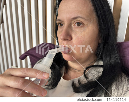Adult woman using a home nebulizer inhaler while sitting in a cozy bedroom, respiratory treatment at home for breathing relief, healthcare and medical therapy concept, realistic lifestyle scene 134119214