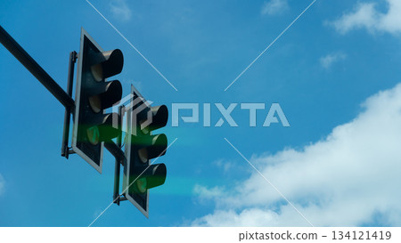 Look up below shows the sides of a double traffic light. Shows the green lights appearing on both sides. Signals a way through. Under blue sky and white clouds at day time. 134121419