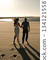 Couple walking barefoot on sandy beach near ocean at sunset, wind blowing her hair in a peaceful romantic scene. 134121558
