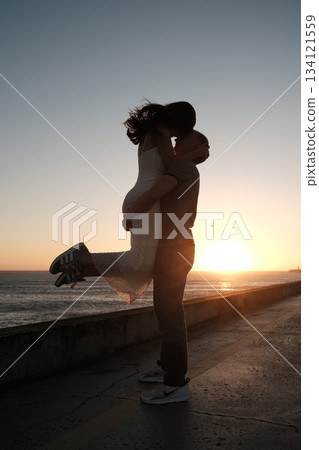Man lifting woman in joyful embrace by the sea at sunset, romantic silhouette moment filled with love and happiness. 134121559