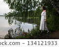 Elegance by the Lake: Woman in White Off-Shoulder Dress Among Birch Trees 134121933