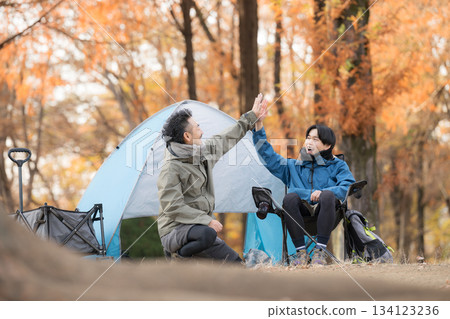 Outdoor holiday scene of two smiling men posing in front of a tent at an autumn campsite, having fun high-fiving 134123236