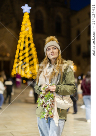 Girl with cone and fir brunch garland stands in middle fair decorated for New Year holidays Girl with cone and fir brunch garland stands in middle fair decorated for New Year holidays 134124411