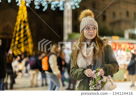 Girl with cone and fir brunch garland stands in middle fair decorated for New Year holidays Girl with cone and fir brunch garland stands in middle fair decorated for New Year holidays 134124416