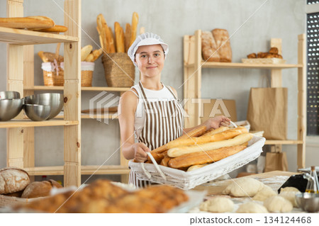 Young girl baker are standing near product range in bakery kitchen with basket loaf of baguettes Young girl baker are standing near product range in bakery kitchen with basket loaf of baguettes 134124468