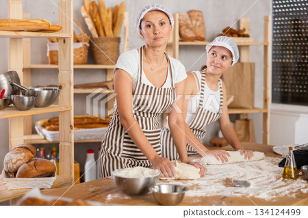 Two skilled female bakers in uniform kneading dough and rolling out dough into sausages before baking in bakehouse 134124499