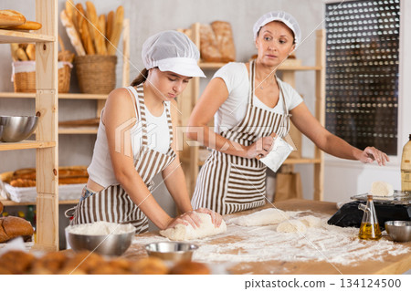 Girl helps an experienced baker prepare dough in kitchen of bakery - together they knead dough, cut it with a knife and weigh it on scales 134124500