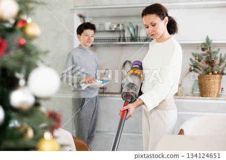 Preparing for Christmas - mother vacuuming floor while a teenage son washes dishes in kitchen Preparing for Christmas - mother vacuuming floor while a teenage son washes dishes in kitchen 134124651
