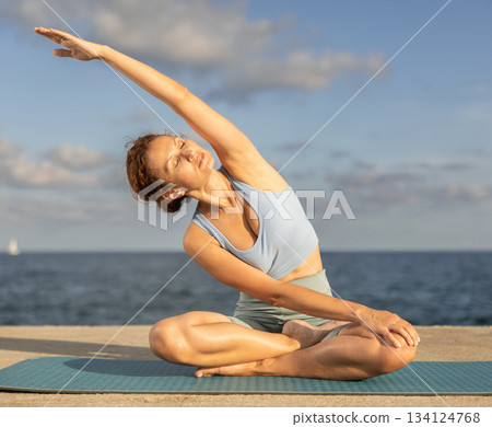 Female fitness trainer performing yoga parsvottanasana sitting on mat on beach 134124768