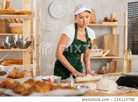 Young woman working at bakery rolling out dough into sausages, female baker kneading dough and shaping baguettes on countertop in kitchen Young woman working at bakery rolling out dough into sausages, female baker kneading dough and shaping baguettes on countertop in kitchen 134124786