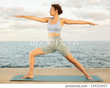 Yoga practice on embankment, female athlete fulfil carry out warrior pose, Virabhadrasana exercise Yoga practice on embankment, female athlete fulfil carry out warrior pose, Virabhadrasana exercise 134124857